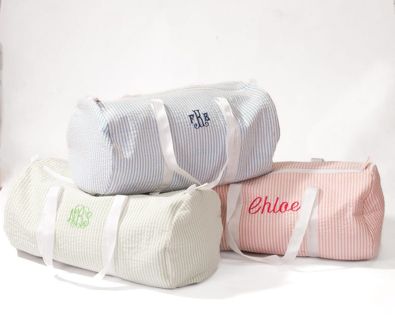 Puede incluir: Tres bolsas de lona a rayas con correas blancas. Las bolsas est&aacute;n personalizadas con los nombres "F.R.H." en azul marino, "Chloe" en rosa y un monograma verde.