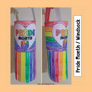 Puede incluir: Una manga de viento cilíndrica de papel decorada con rayas de arcoíris hechas con ceras. Presenta un gran corazón blanco con el texto PRIDE MONTH y un gráfico de un puño en alto. Un asa de papel blanco está unida a la parte superior.