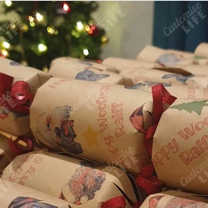 Op de afbeelding: Close-up van bruine kerstcrackers, elk versierd met rode linten en hondenillustraties. De crackers hebben de tekst "Happy Woofmas Ralff". Op de achtergrond staat een kerstboom met vage lichten.