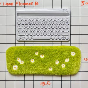 May include: A white wireless keyboard sits above a rectangular green item resembling a lawn, adorned with small white flowers. The green item measures 15.5 inches long and 6 inches wide. The image is set against a grid background.
