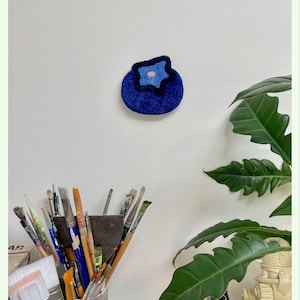 May include: A circular, tufted wall hanging shaped like a blueberry, featuring deep blue yarn with a lighter blue and pale pink center. The text "tactile art" is printed in a small blue rectangle in the upper left corner of the composition.