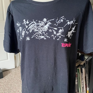May include: Black t-shirt featuring a white graphic of skeletons riding a rooster and other figures. The word "ESPION" is printed in pink below the graphic. The shirt is displayed on a mannequin.