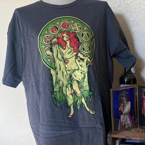 Peut inclure: T-shirt gris foncé avec un motif d'une femme zombie aux cheveux roux et vêtue de vert et de jaune. Le motif est encadré dans un cercle vert et doré. Le t-shirt est présenté sur un mannequin.