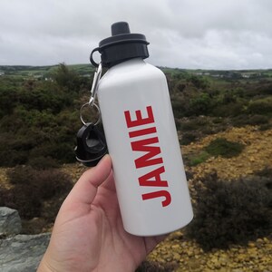 K&ouml;nnte beinhalten: Eine wei&szlig;e Wasserflasche mit schwarzem Deckel und dem Namen "JAMIE" in rot aufgedruckt.