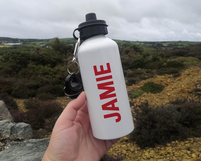 K&ouml;nnte beinhalten: Eine wei&szlig;e Wasserflasche mit schwarzem Deckel und dem Namen "JAMIE" in rot aufgedruckt.