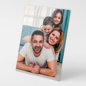 May include: A family of four pose for a photograph. The father is lying on the floor with his two children on his back. The mother is standing behind them, smiling. They are all looking at the camera.