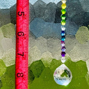 Op de afbeelding: Een regenboogkleurige kristallen zonnevanger, opgehangen aan een transparante draad. De zonnevanger heeft een reeks kleurrijke kralen en een grote kristallen bol. Een rode liniaal, die tot 23 cm meet, staat in de afbeelding.
