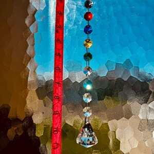 May include: A decorative crystal prism suncatcher, approximately 33 cm long, hangs from a metal hook. The prism is composed of clear, faceted crystals and colourful beads in a rainbow spectrum. The background is a textured glass window.
