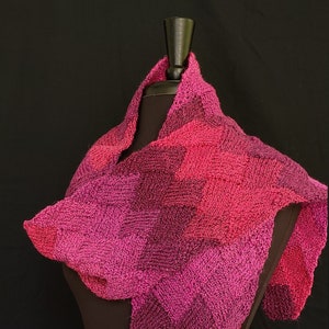 May include: A pink and purple knitted scarf with a chevron pattern. The scarf is draped over a black mannequin.
