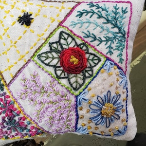 May include: A small, square, embroidered pincushion with a patchwork design. The cushion features various floral and geometric patterns in shades of red, green, blue, yellow, and purple. The pincushion is likely handmade and sits on a vintage sewing machine.