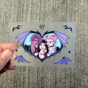 May include: A rectangular clear sticker featuring a heart-shaped frame with purple bat wings. Inside the heart are three illustrated characters with pink and dark hair. The text HEX is printed above the heart, accompanied by four small star and wing accents.