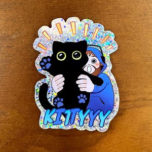 Könnte beinhalten: Ein holografischer Aufkleber mit einer schwarzen Katze, die von einer Person in einem blauen Kapuzenpullover gehalten wird. Die Katze hat gelbe Augen und blaue Pfotenabdrücke. Das Wort "KITTYYY" steht unten in Blau.