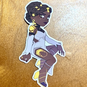 May include: A sticker of a cartoon woman with brown skin, wearing a white dress with gold accents and yellow boots. She has a gold crown and gold jewelry. Her hair is styled in braids with gold accents.