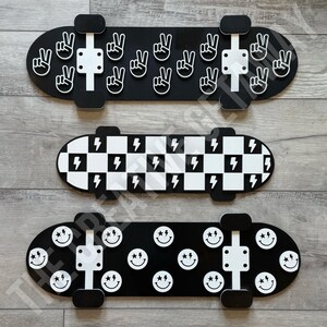 Op de afbeelding: Drie decoratieve skateboards. De bovenste is zwart met witte vredestekens. De middelste heeft een zwart-wit geblokt patroon met bliksemschichten. De onderste is zwart met witte smiley gezichten.