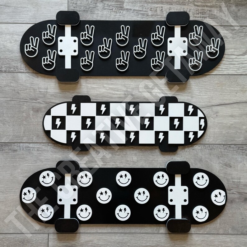 Op de afbeelding: Drie decoratieve skateboards. De bovenste is zwart met witte vredestekens. De middelste heeft een zwart-wit geblokt patroon met bliksemschichten. De onderste is zwart met witte smiley gezichten.