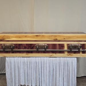 May include: A wooden casket with a natural wood finish, featuring a combination of light and dark wood tones. The casket has metal handles and decorative accents. The casket is displayed on a white draped surface.