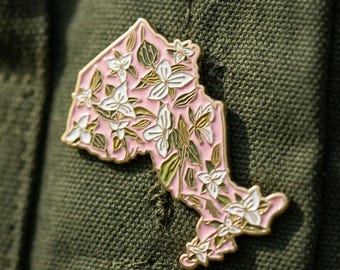 May include: A pink enamel pin in the shape of the Canadian province of Ontario, featuring white flowers and green leaves. The pin has a gold border and is set against a green fabric background.