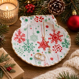 May include: A white shell-shaped trinket dish with a gold-painted scalloped edge, featuring a festive pattern of red and green snowflakes, dots, and hanging lines. A delicate gold chain necklace and two gold rings rest on the dish surface.