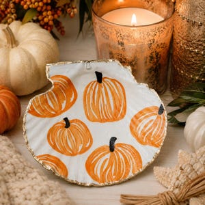 May include: A white scallop shell dish featuring hand-painted orange pumpkins with black stems. The edges of the shell are trimmed in a metallic gold finish. The surface of the shell is smooth and curved, displaying five distinct pumpkin illustrations.