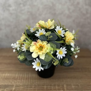 May include: A floral arrangement with yellow and white daisies, yellow flowers, and green foliage. The flowers are in a black pot, creating a vibrant display. The arrangement is set on a wooden surface.