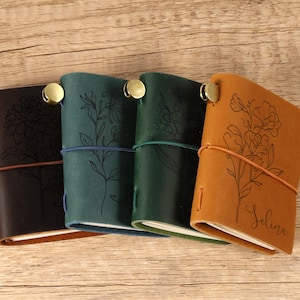 Könnte beinhalten: Vier Lederjournale in Braun-, Türkis- und Grüntönen, jedes mit einem Blumendesign und einem Messingverschluss. Das Journal rechts ist hellbraun und trägt den eingravierten Namen "Selina".