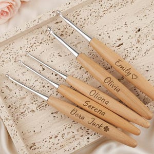 May include: A collection of personalized crochet hooks with wooden handles and silver metal tips. Each hook is engraved with a name: Emily, Olivia, Oliona, Serena, and Dora Jack. The hooks are arranged on a textured tray with a floral arrangement.