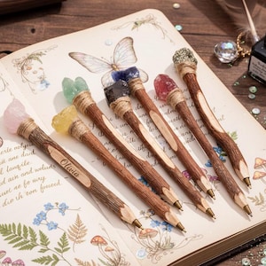 Può includere: Una collezione di penne decorative con top di pietre preziose, esposte su un diario aperto con illustrazioni floreali e di farfalle. Le penne hanno fusti in legno e sono sormontate da cristalli di vari colori. Una penna è incisa con il nome "Olivia".