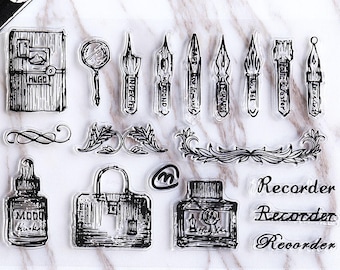 May include: Clear stamp set featuring various vintage-style illustrations. Includes a journal, magnifying glass, ink bottles, pens, a briefcase, and decorative flourishes. The word "Recorder" is written three times in a script font.