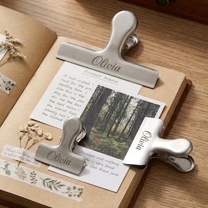 May include: Three silver metal binder clips engraved with the name "Olivia" on a notebook with handwritten notes and a photograph. The notebook is open, with dried flowers and decorative tape.