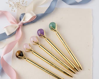 May include: Five gold pens with gemstone tops in various colors: green, purple, pink, and brown. The pens are arranged on a textured paper surface, with a ribbon bow in the background. The pens have the date 02/02/2026 engraved on them.