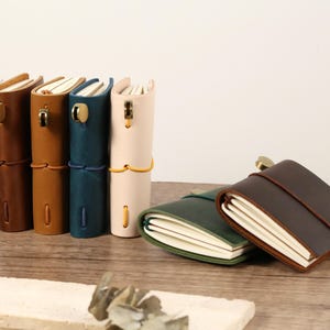 May include: A collection of leather-bound journals in various colors, including brown, tan, teal, and cream. The journals are standing upright, with a few lying flat. Each journal has a clasp or tie closure.