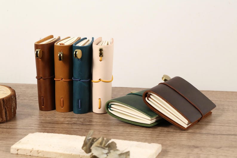 May include: A collection of leather-bound journals in various colors, including brown, tan, teal, and cream. The journals are standing upright, with a few lying flat. Each journal has a clasp or tie closure.