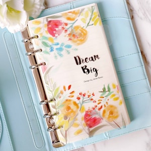 May include: A light blue leather bound planner with a floral design and the text "Dream Big" on the cover. The planner has a silver ring binder.