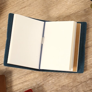 May include: An open, blue journal with blank white pages and a blue elastic closure. The journal is on a wooden surface, with a few brown and black objects in the background.