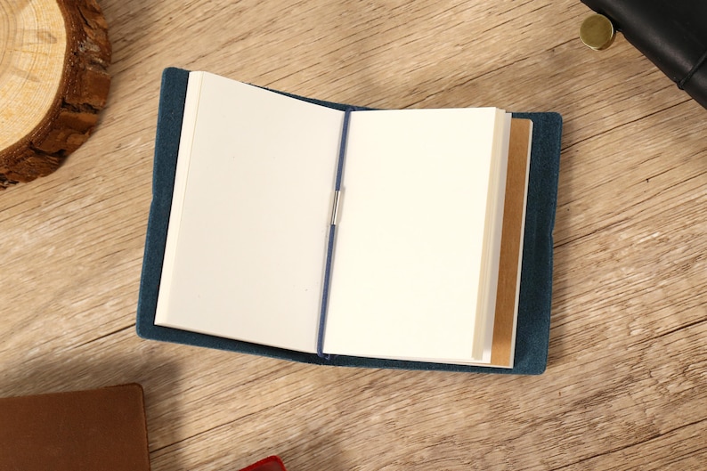 May include: An open, blue journal with blank white pages and a blue elastic closure. The journal is on a wooden surface, with a few brown and black objects in the background.