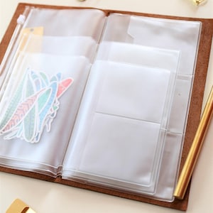 May include: A brown leather bound journal with clear plastic pockets for holding papers, stickers, and other small items. The journal is open and the pockets are empty.