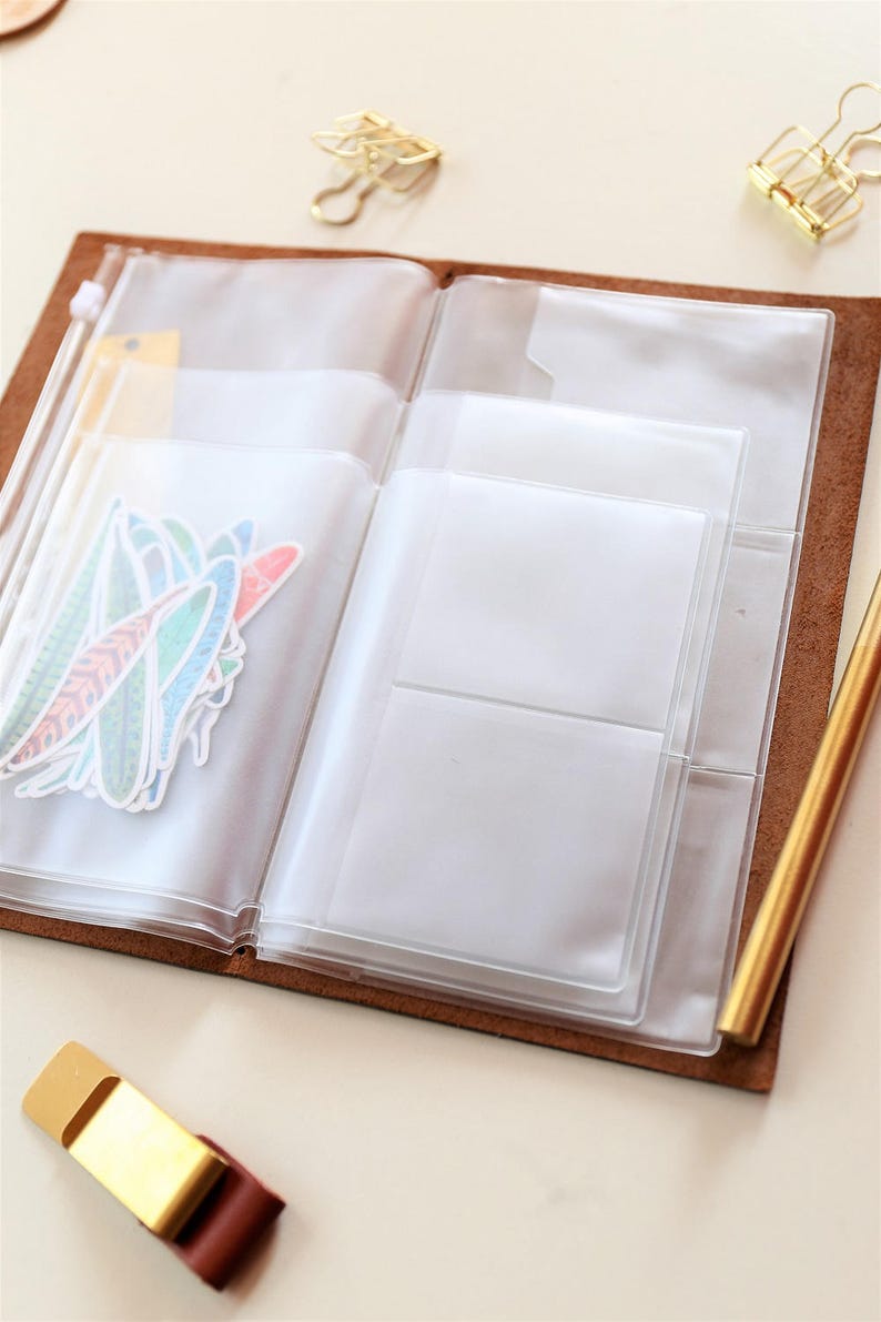 May include: A brown leather bound journal with clear plastic pockets for holding papers, stickers, and other small items. The journal is open and the pockets are empty.