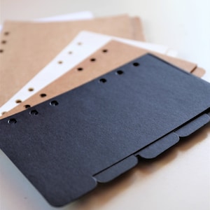 Op de afbeelding: Een zwarte scheider met tabbladen voor een planner of notitieboek. De scheider ligt op bruin en wit papier met gaten.