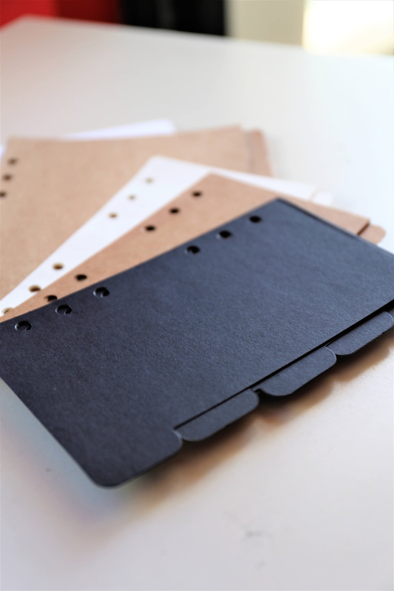 Op de afbeelding: Een zwarte scheider met tabbladen voor een planner of notitieboek. De scheider ligt op bruin en wit papier met gaten.