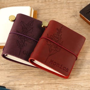 May include: Close-up of several leather journals in shades of burgundy and brown, with floral and botanical engravings. One journal is open, revealing blank pages. The journals are secured with elastic bands and brass closures. The word "Monica" is engraved on one journal.
