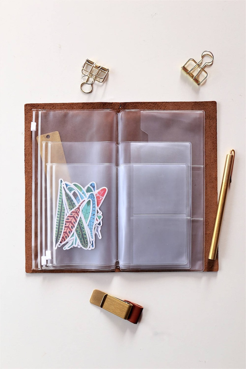 May include: A brown leather notebook with clear plastic pockets and zippered pouches. The notebook is open and has a variety of stickers and a ruler inside. There are also two gold metal clips and a gold pen on the white surface.