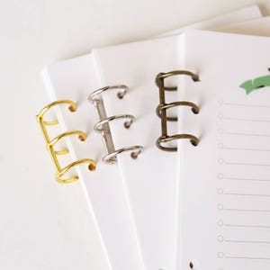 Puede incluir: Tres anillos de encuadernación metálicos en dorado, plateado y bronce. Los anillos están diseñados para un cuaderno o una agenda.