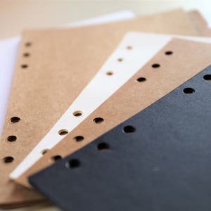 Op de afbeelding: Een stapel van drie verschillende gekleurde papieren notitieboekjes met geperforeerde gaten langs de zijkant. De notitieboekjes zijn wit, bruin en zwart.