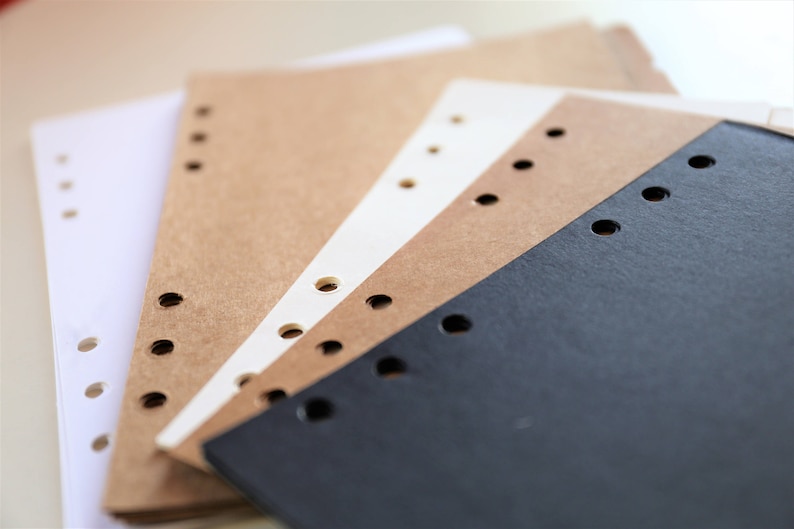 Op de afbeelding: Een stapel van drie verschillende gekleurde papieren notitieboekjes met geperforeerde gaten langs de zijkant. De notitieboekjes zijn wit, bruin en zwart.