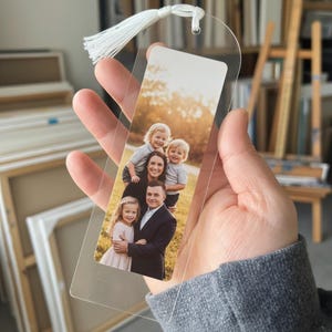 Peut inclure: Marque-page en acrylique transparent avec une photo de famille et un pompon blanc. La photo rectangulaire montre une famille de cinq personnes, avec deux jeunes garçons, une fille et leurs parents. Le marque-page est tenu par une personne.
