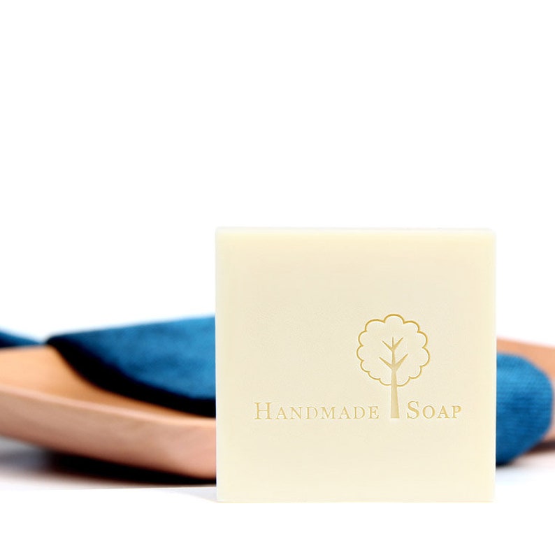 Puede incluir: Una barra de jab&oacute;n artesanal blanca con un logotipo de &aacute;rbol y las palabras "Handmade Soap" impresas en la parte delantera.