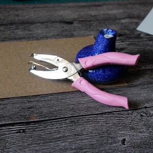 Puede incluir: Una perforadora plateada con mangos rosas descansa sobre una superficie marr&oacute;n, junto a un peque&ntilde;o jarr&oacute;n de cer&aacute;mica azul. La perforadora est&aacute; abierta. Los mangos son de color rosa.