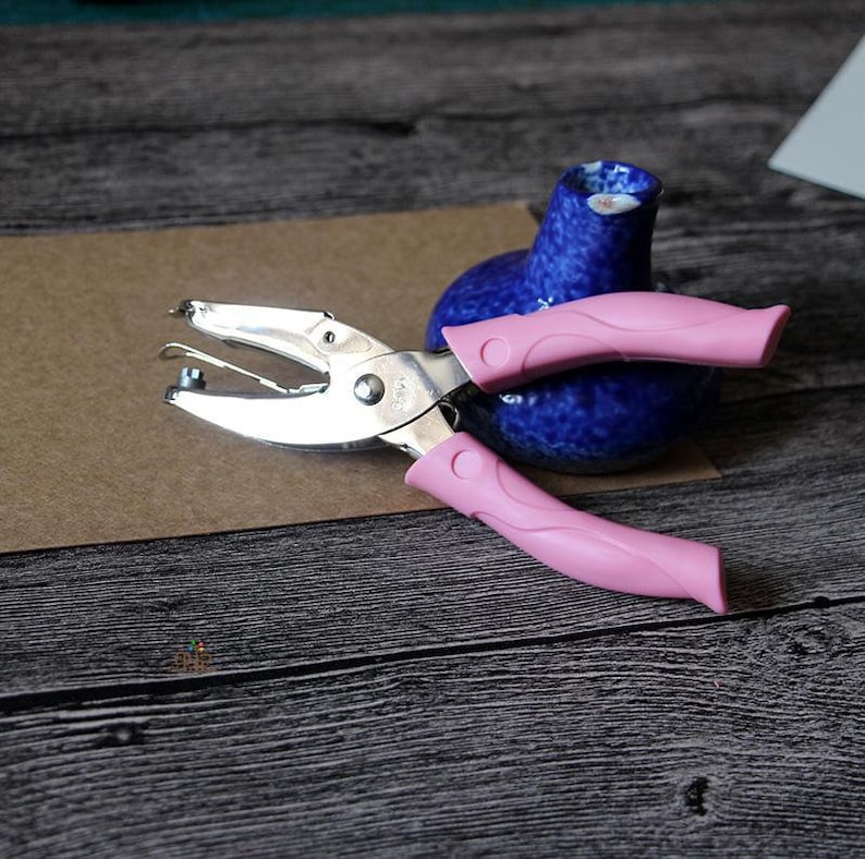 Puede incluir: Una perforadora plateada con mangos rosas descansa sobre una superficie marr&oacute;n, junto a un peque&ntilde;o jarr&oacute;n de cer&aacute;mica azul. La perforadora est&aacute; abierta. Los mangos son de color rosa.