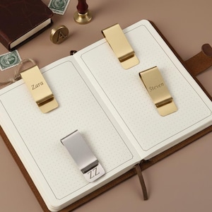 Puede incluir: Un cuaderno abierto muestra cuatro clips de marcapáginas metálicos. Tres son dorados y uno plateado. Los clips dorados están grabados con los nombres "Zara" y "Steven". El clip plateado está grabado con "ZZ".