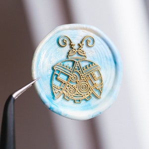 Könnte beinhalten: Ein blaues und weißes Wachssiegel mit einem goldenen Design eines stilisierten Käfers mit Zahnrädern und Rädern. Das Siegel wird von einer Pinzette gehalten.